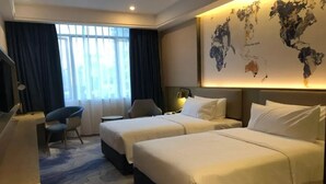 A hotel room with two beds, a desk, a chair, a world map on the wall, and a large window with curtains.