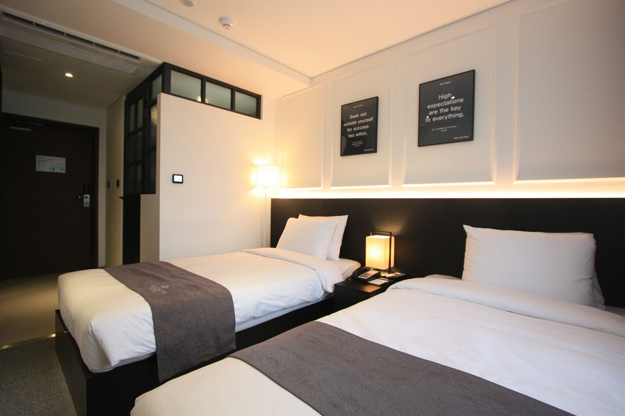 Habitació d'hotel amb dos llits, una tauleta de nit amb una làmpada i pòsters motivacionals a la paret.