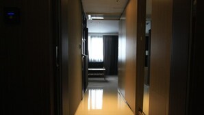 A hotel hallway with a wooden door on the right, a bench in the middle, and a window with curtains at the end. - Suwon Vella Suite Hotel (Suwon)