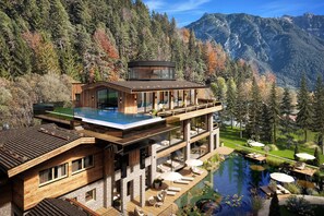 Een modern huis met een glazen overloopzwembad, een houten terras en een berg op de achtergrond.