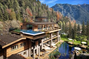 Una casa moderna con piscina infinita de vidrio, terraza de madera y montañas al fondo.