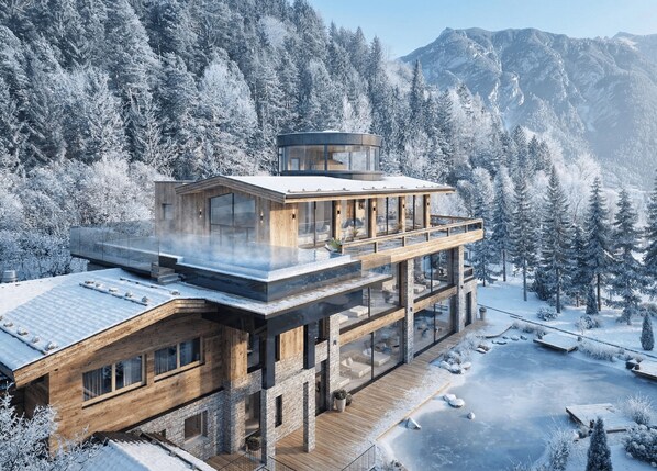 Un edificio moderno de varios pisos con fachada de vidrio, terraza de madera y vista a un bosque nevado.
