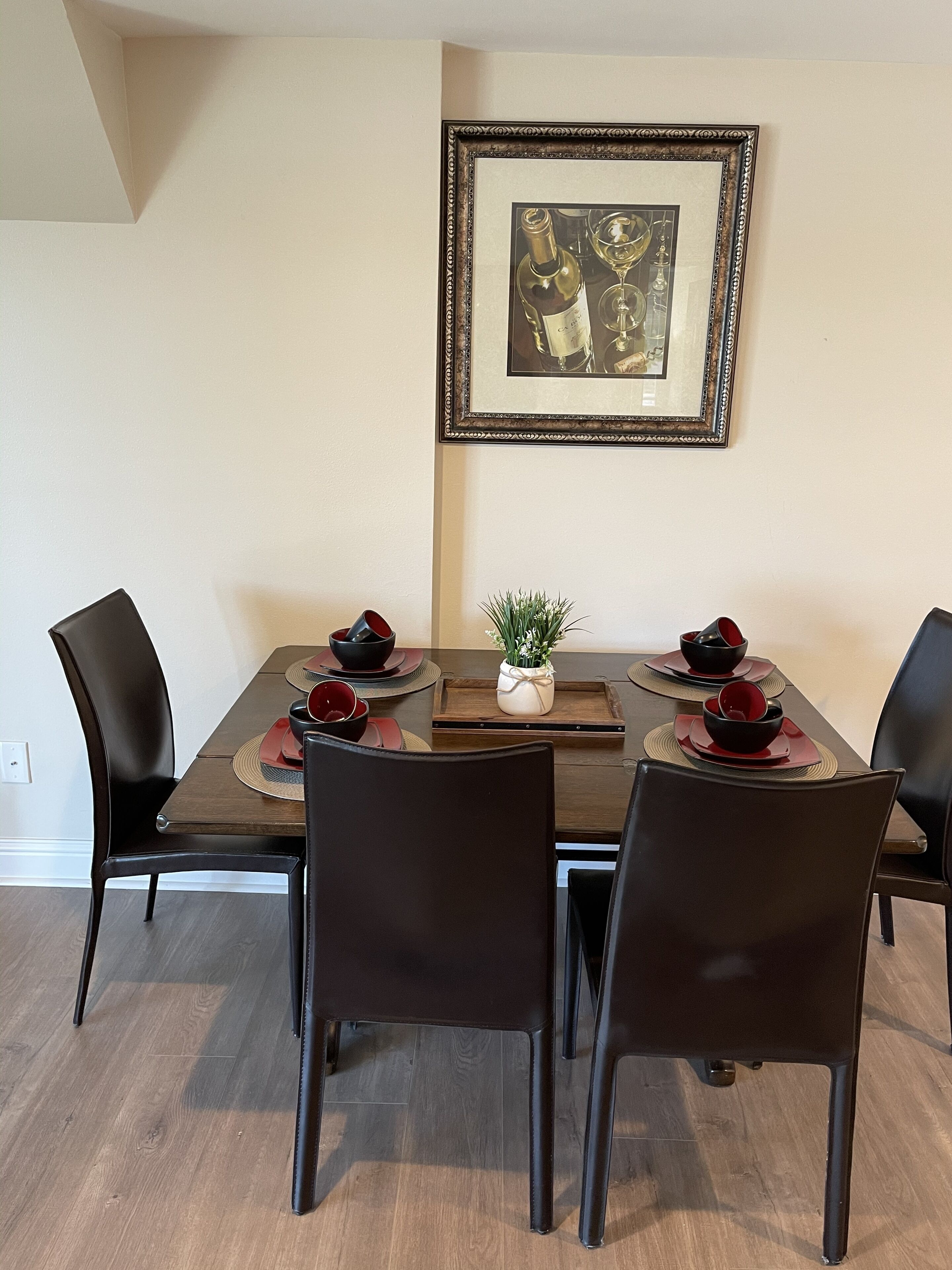 A dining area with a rectangular table set for four, surrounded by black chairs. A framed picture hangs on the wall above the table.