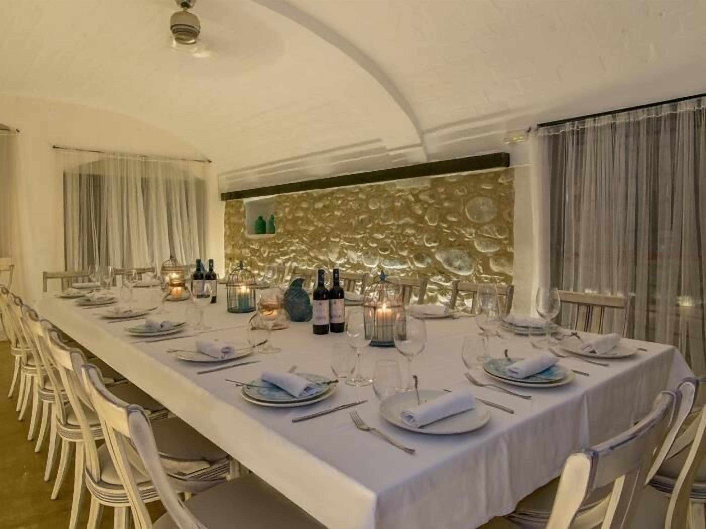 Une longue table à manger, recouverte de nappes blanches, agrémentée d’assiettes et de bouteilles de vin, et entourée de chaises blanches.