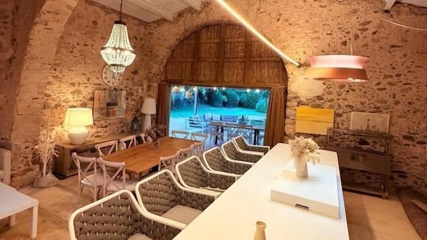 Um interior rústico com paredes de pedra, uma mesa de jantar de madeira, assentos acolchoados e iluminação pendente.