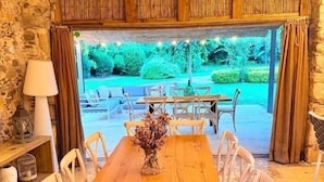 Uma mesa de jantar de madeira com cadeiras brancas, um vaso com flores secas e vista para uma área de estar ao ar livre com uma parede de pedra e teto de madeira.