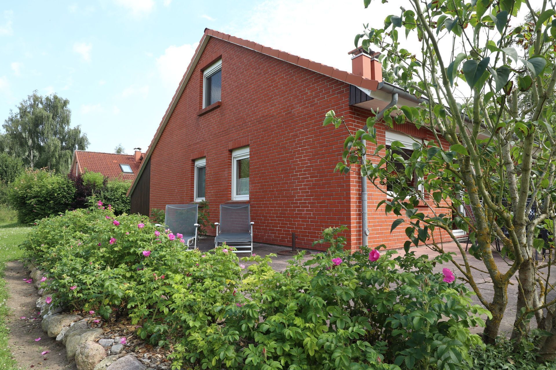 A red brick house with a sloped roof, a front door, and two windows. There is a garden with pink flowers and a tree in the foreground.
