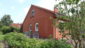 Ein rotes Ziegelhaus mit einem geneigten Dach, einer Haustür und zwei Fenstern. Im Vordergrund befindet sich ein Garten mit rosa Blumen und einem Baum.