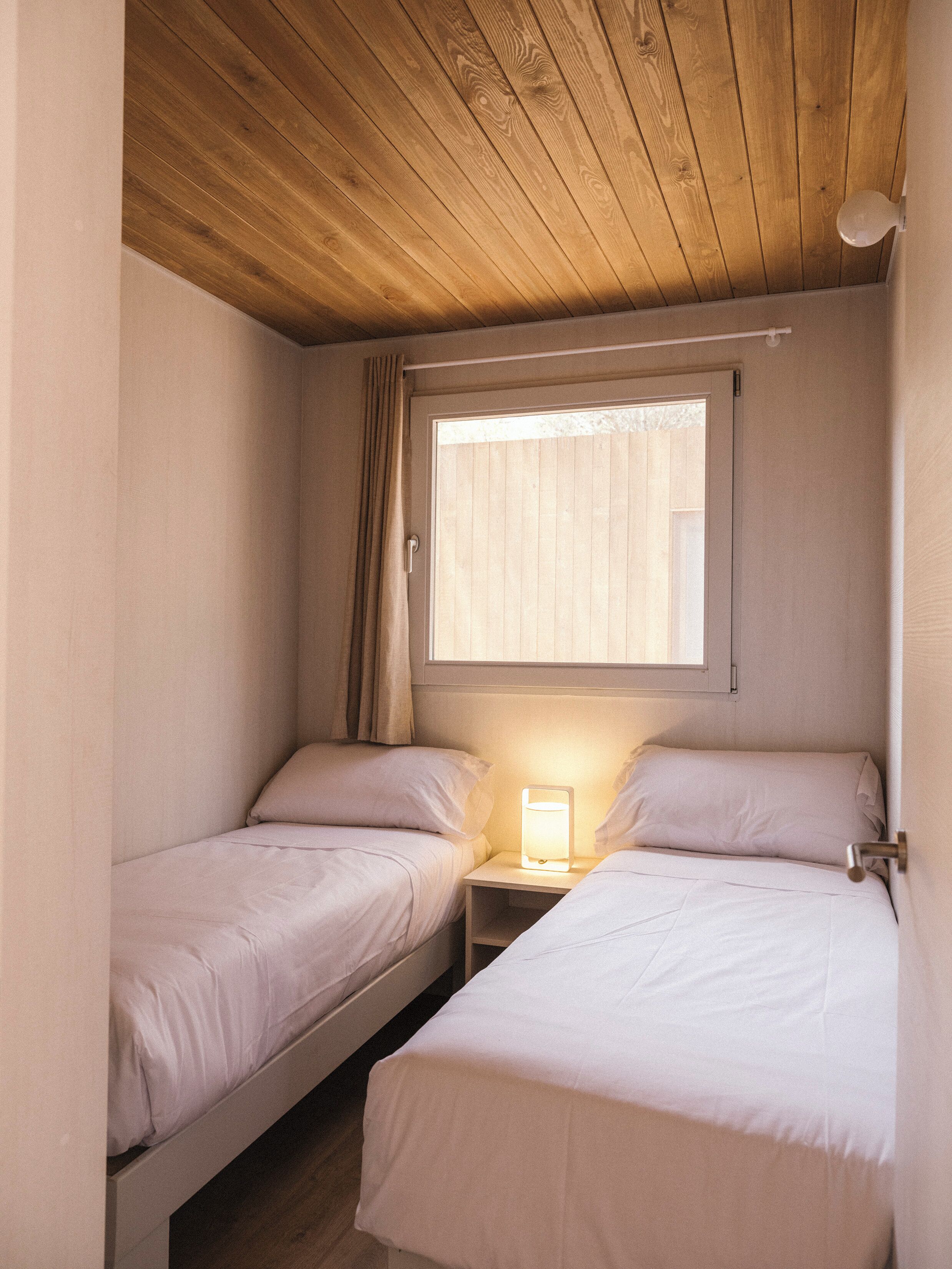 A small, cozy room with two single beds, a nightstand with a lamp, a window with blinds, and a wooden ceiling.