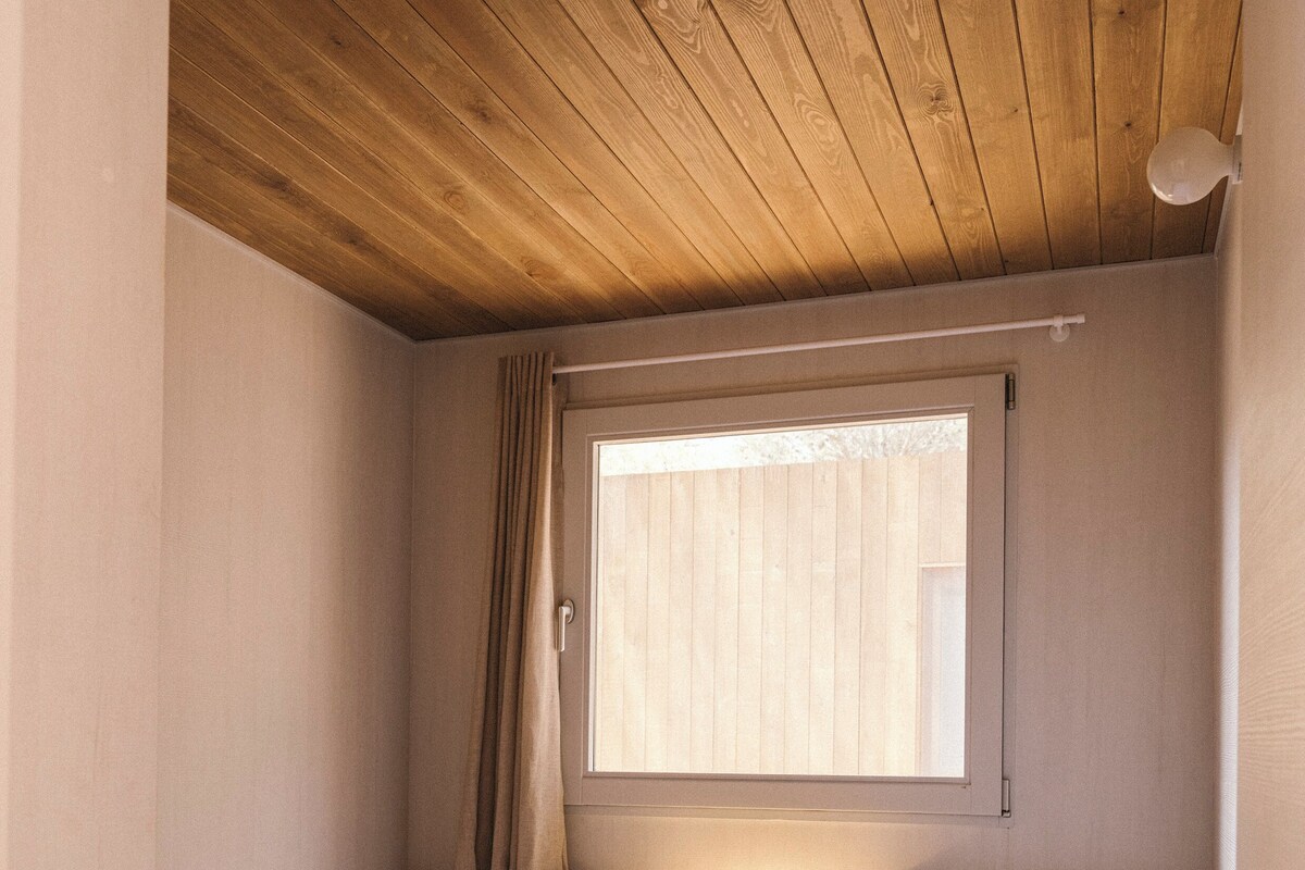 A small, cozy room with two single beds, a nightstand with a lamp, a window with blinds, and a wooden ceiling.