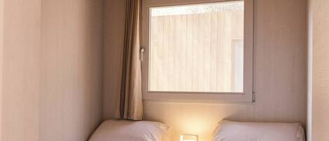 A small, cozy room with two single beds, a nightstand with a lamp, a window with blinds, and a wooden ceiling.