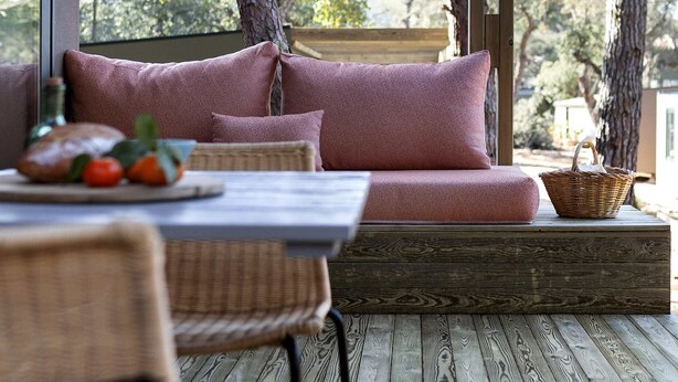 A cozy outdoor seating area with a wooden deck, a cushioned bench, and a wicker table with a bottle and fruit. -  Wecamp Cala Montgó (L'Escala)