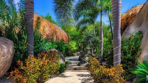 Un sentier qui traverse un jardin tropical, bordé de palmiers, de rochers et agrémenté d’une structure au toit de chaume.