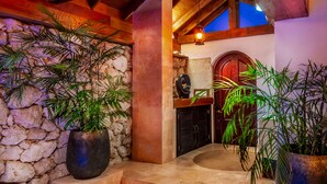Un mur de pierre avec un escalier en bois menant à une porte. De grandes plantes en pot sont disposées de part et d’autre de l’escalier.