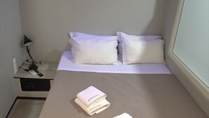 Un lit bien fait, avec du linge de lit blanc et des oreillers, une table de chevet avec une lampe et une étagère fixée au mur avec une télécommande.
