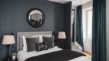 Een moderne slaapkamer met een grijs hoofdeinde, een groot bed met wit en grijs beddengoed, twee bedlampen, een ronde spiegel boven het bed en een raam met turquoise gordijnen.
