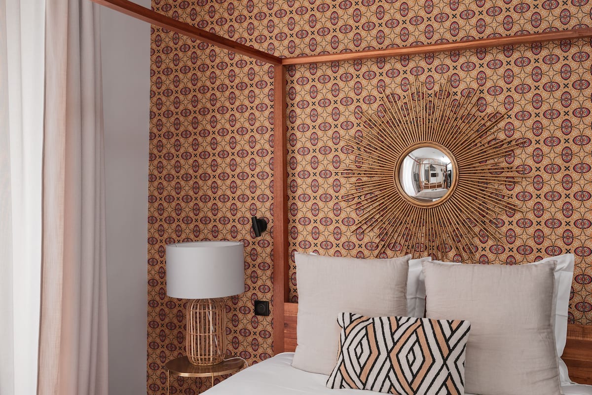 Habitación de hotel con papel tapiz estampado, un espejo redondo, una cama con dos almohadas, una mesita de noche con lámpara y un cabecero de madera.