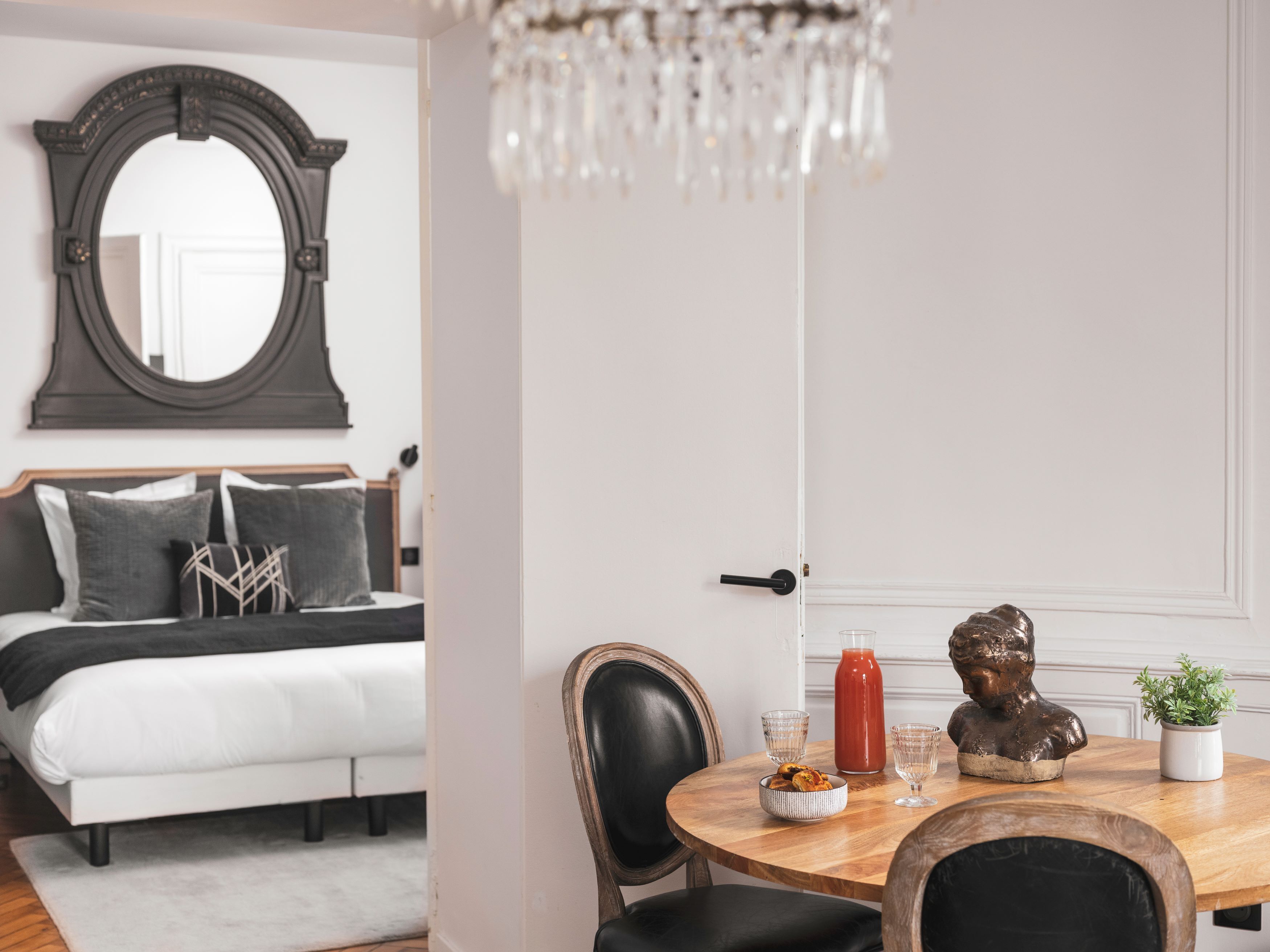 A bedroom with a bed, bedside table, and a mirror. There is a dining area with a table, chairs, and a chandelier.