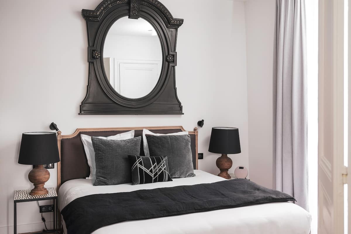 Un dormitorio ordenado con un espejo grande, dos lámparas de noche, una cama con colcha blanco y negro y una mesita de noche con lámpara.