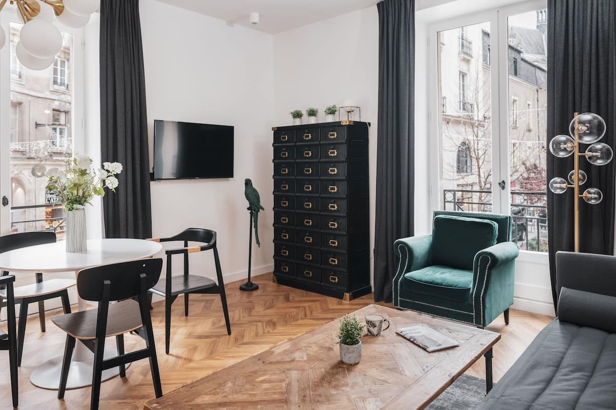 Una sala de estar moderna con piso de madera en espiga, un mueble de almacenamiento negro, un sillón verde, un sofá negro y un comedor con mesa blanca y sillas negras.