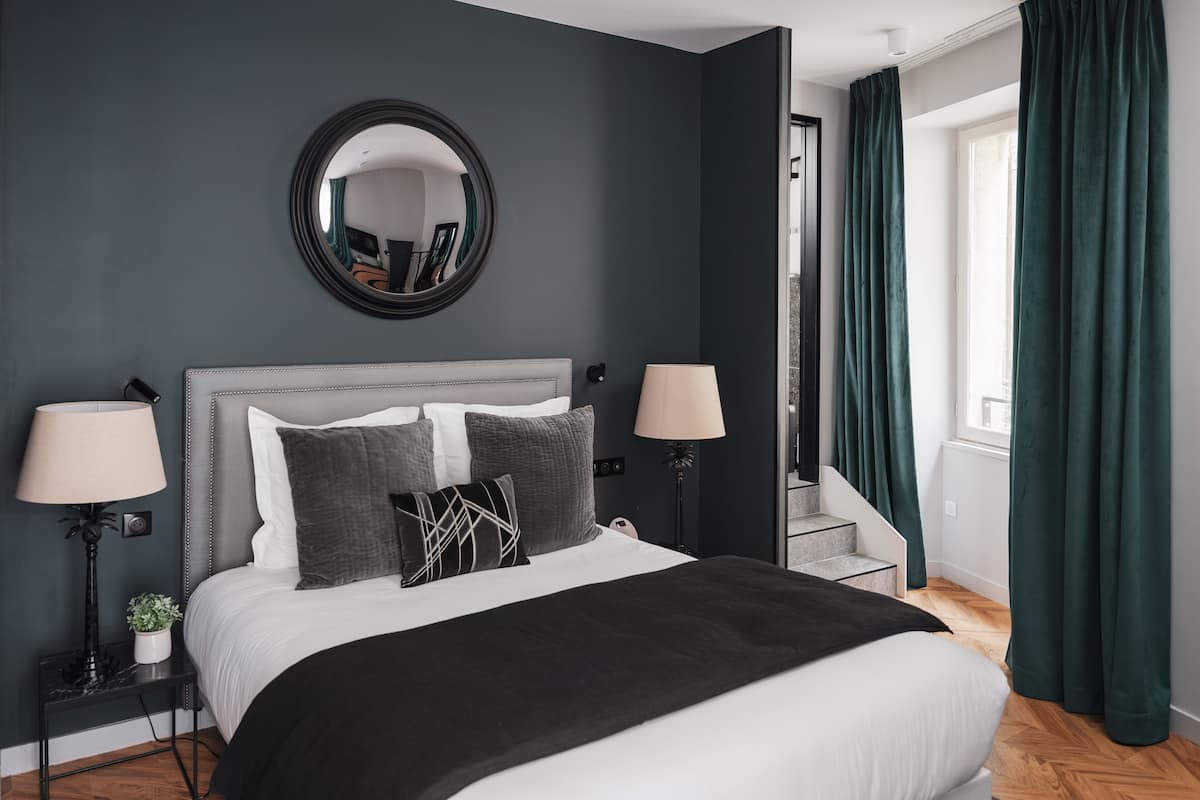 Un dormitorio moderno con un cabecero gris, un espejo redondo, dos lámparas de noche, un plaid negro y vistas de una escalera.