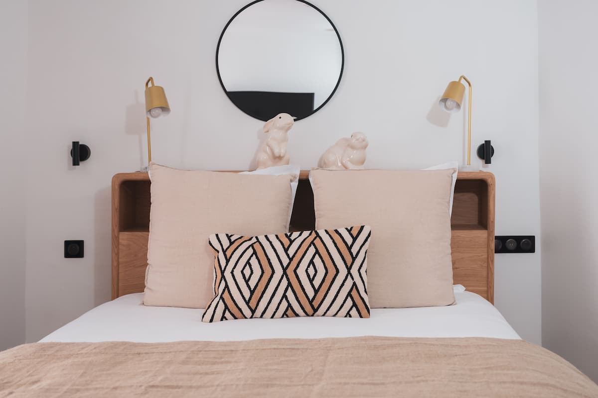 Una cama con cabecera de madera, dos lámparas fijadas a la pared, un espejo redondo y dos pequeñas figuras decorativas en la cabecera.