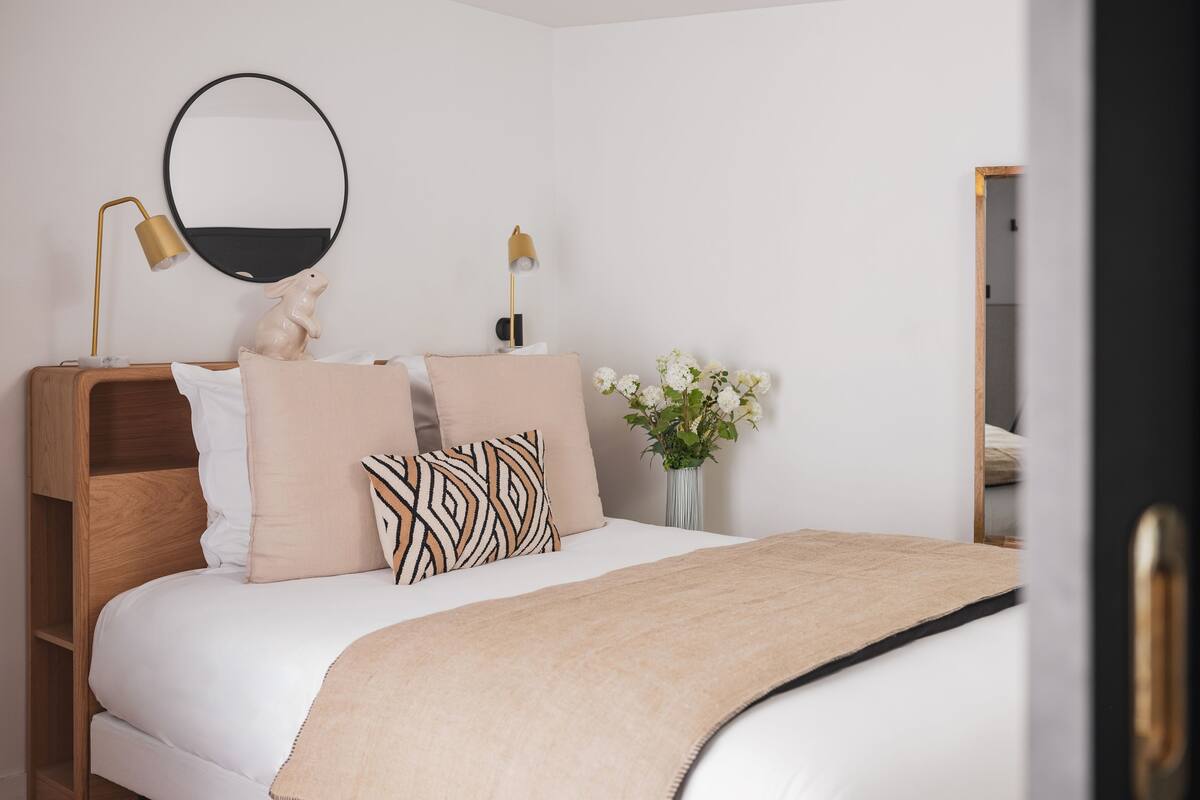 Un dormitorio con cama, lámparas de noche, un espejo, cómoda y un florero con flores.