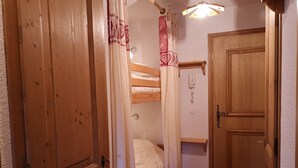 Un dormitorio con puerta de madera, mesita de noche, cama con ropa blanca, cortina y teléfono fijo en la pared.