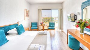 A hotel room with a bed, two chairs, a small table with flowers, a TV, and a balcony with a view. - Paphos Gardens Apartments (Paphos)