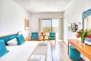 Habitación de hotel con cama, dos sillas, una mesita con flores, televisor y balcón con vistas.