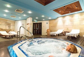 A hot tub with a person relaxing, lounge chairs, a wooden ceiling, and a tiled wall. - Paphos Gardens Apartments (Paphos)