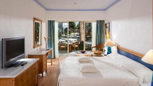 A hotel room with a bed, a TV, a desk, and a view of the pool area. - Paphos Gardens Apartments (Paphos)