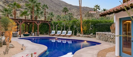 A swimming pool with lounge chairs, surrounded by palm trees and a mountainous backdrop.