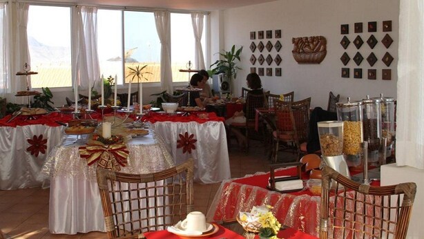 Un comedor con mesas dispuestas para una comida, con manteles blancos y rojos, sillas de mimbre y vistas al exterior a través de amplios ventanales.