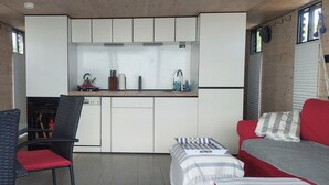 Une cuisine compacte avec des meubles blancs, un petit plan de travail et un évier. On y trouve une chaise rouge et un canapé à rayures dans l’espace salon.