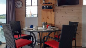 Un intérieur en bois avec une table ronde, quatre chaises, une horloge, un téléviseur et une fenêtre donnant sur un bâtiment situé au loin.