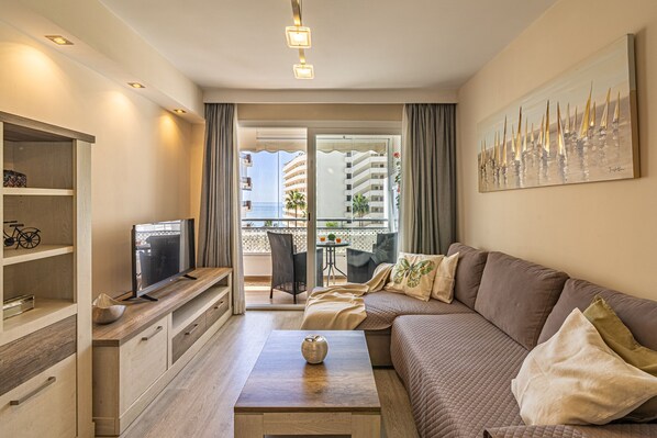 A modern living room with a sofa, a coffee table, a TV, and a balcony with a view of buildings.