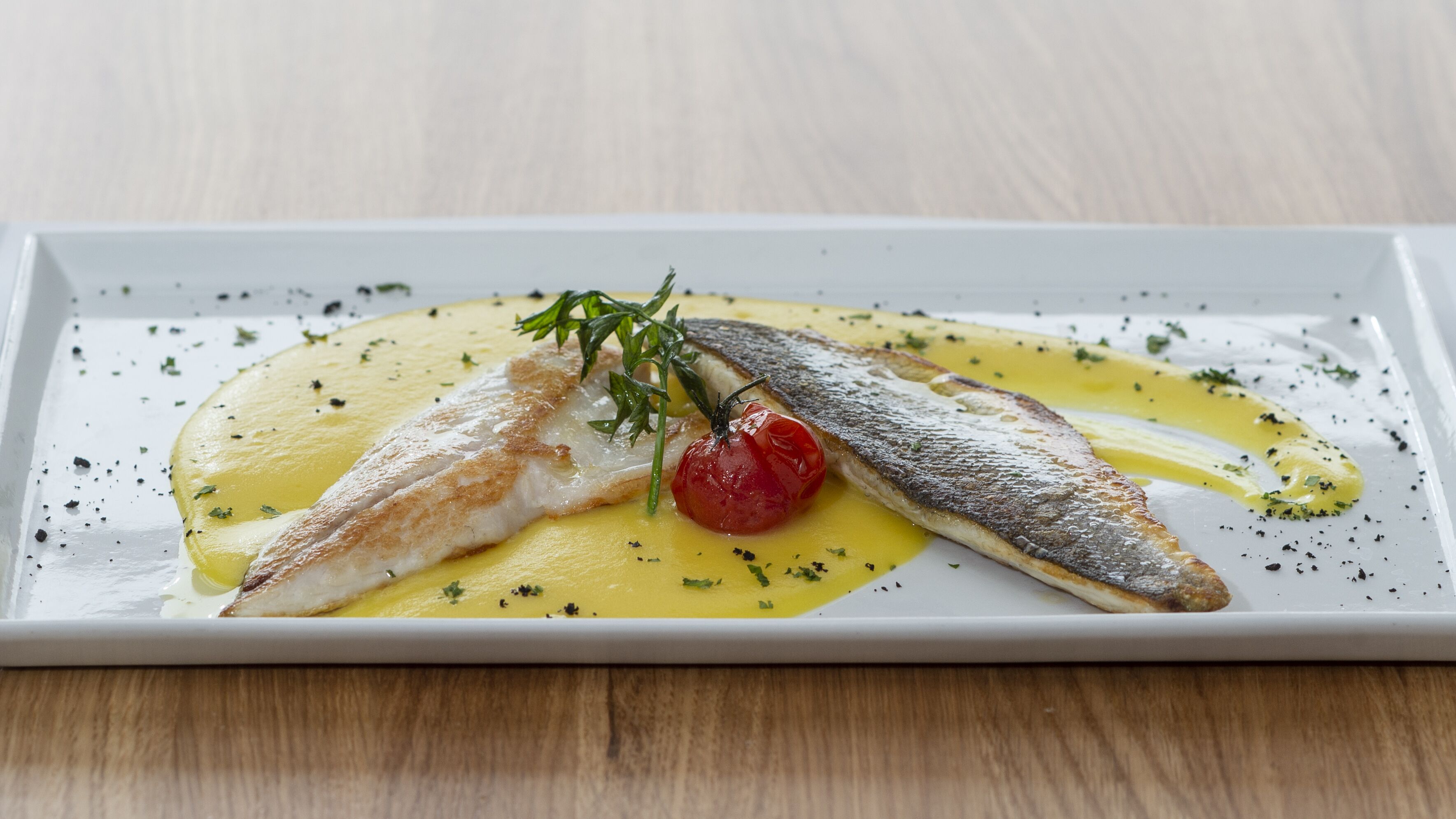 Une assiette rectangulaire blanche sur laquelle on a disposé un morceau de poisson, une tomate cerise et une garniture, le tout nappé d’une sauce jaune.