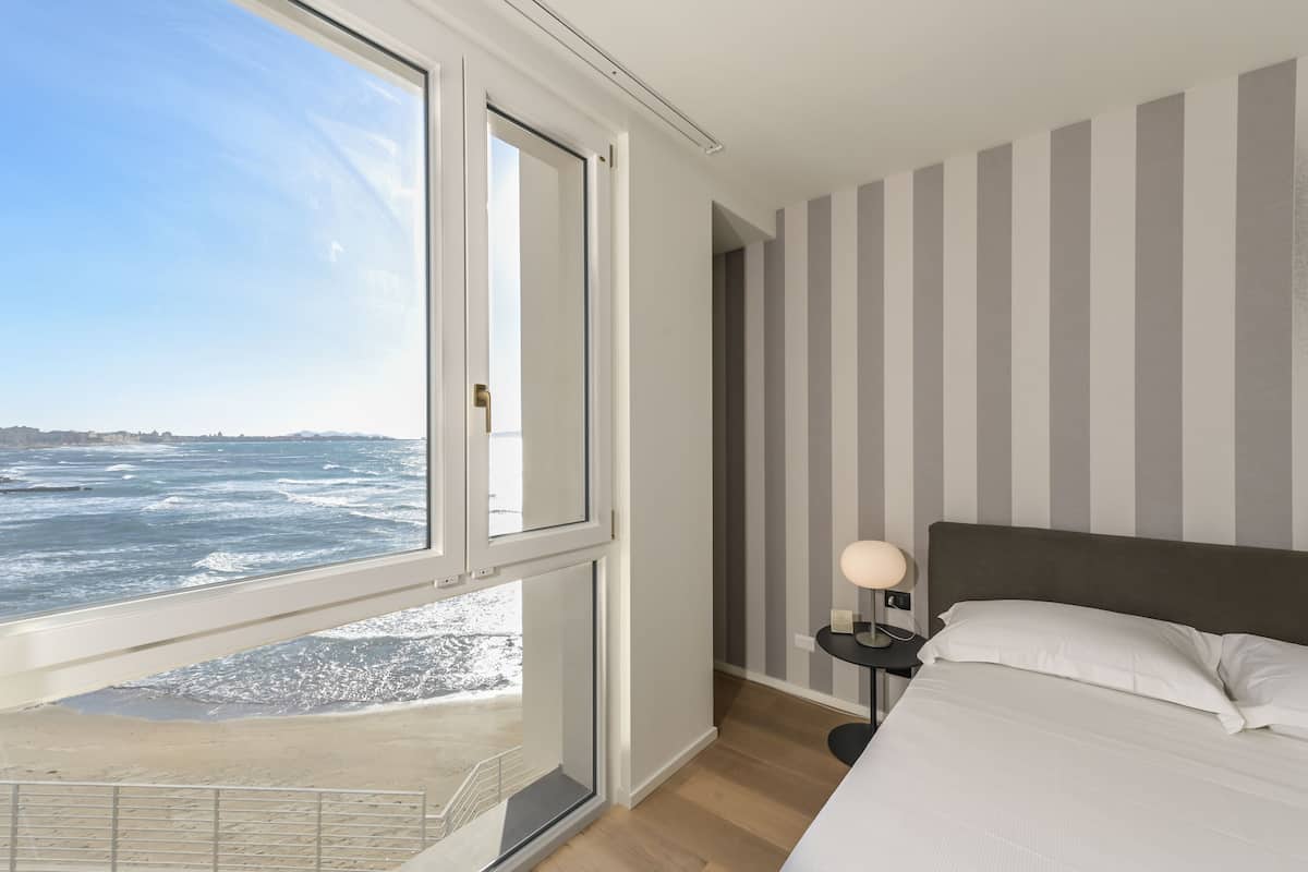 Une chambre avec une grande fenêtre donnant sur l’océan, un lit avec une literie blanche, une table de chevet avec une lampe et des murs à rayures.