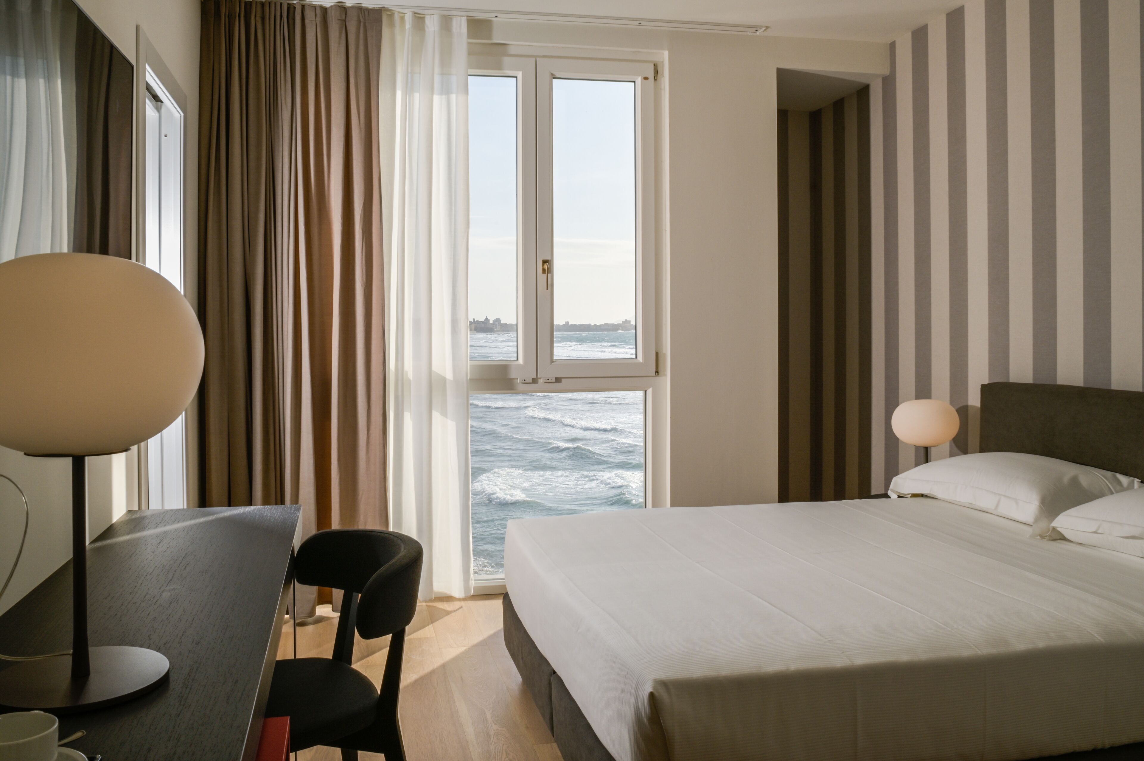 Een hotelkamer met een bed, een bureau, een stoel en uitzicht op zee door een raam.