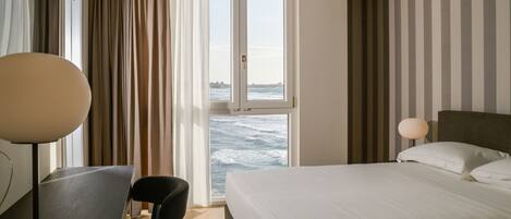 Een hotelkamer met een bed, een bureau, een stoel en uitzicht op zee door een raam.