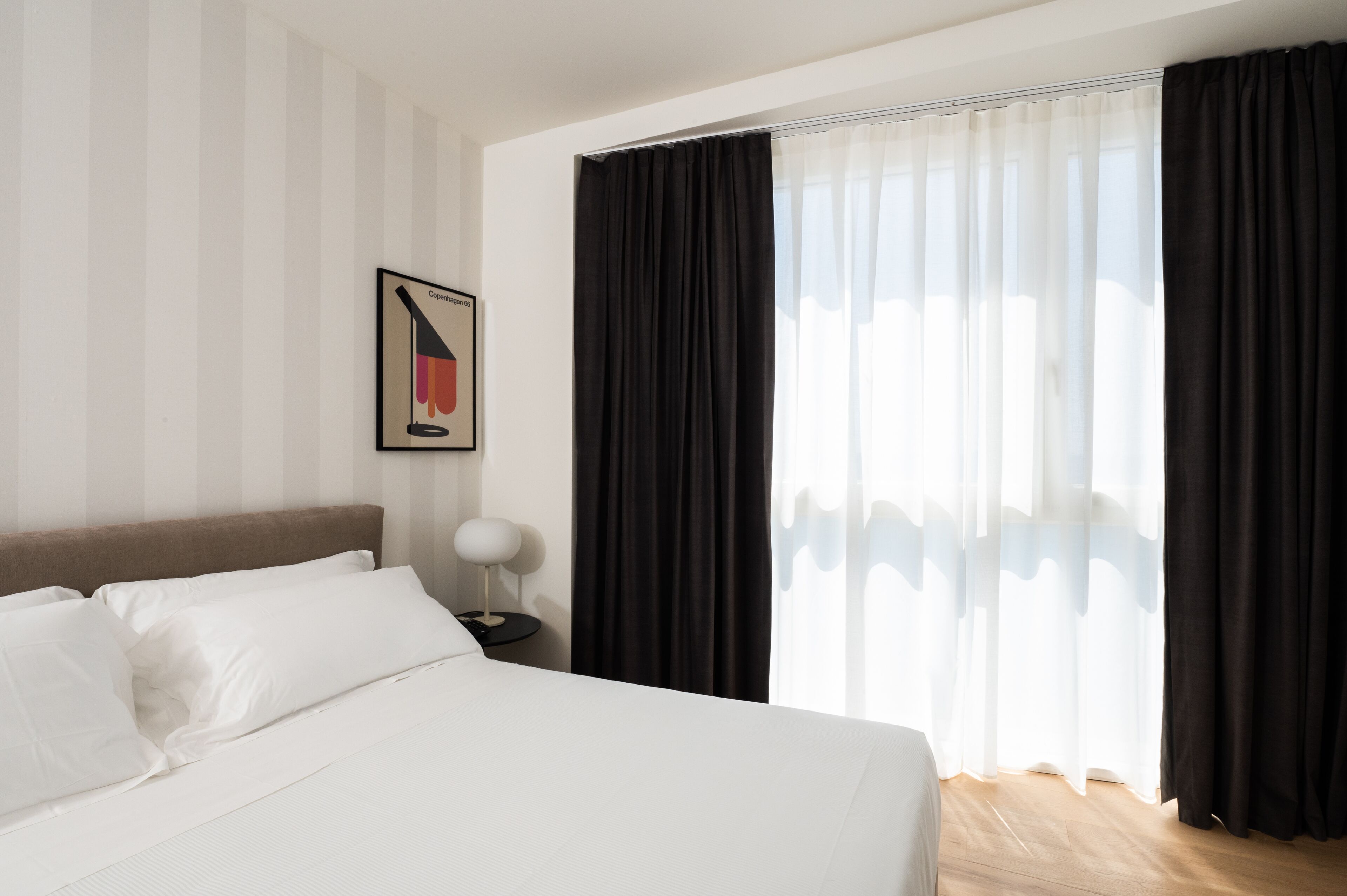 A hotel room with a bed, a nightstand, a lamp, and a window with black curtains.