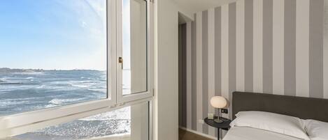 Un dormitorio con una ventana grande que da al océano, una cama con sábanas blancas, una mesita de noche con una lámpara y paredes a rayas.