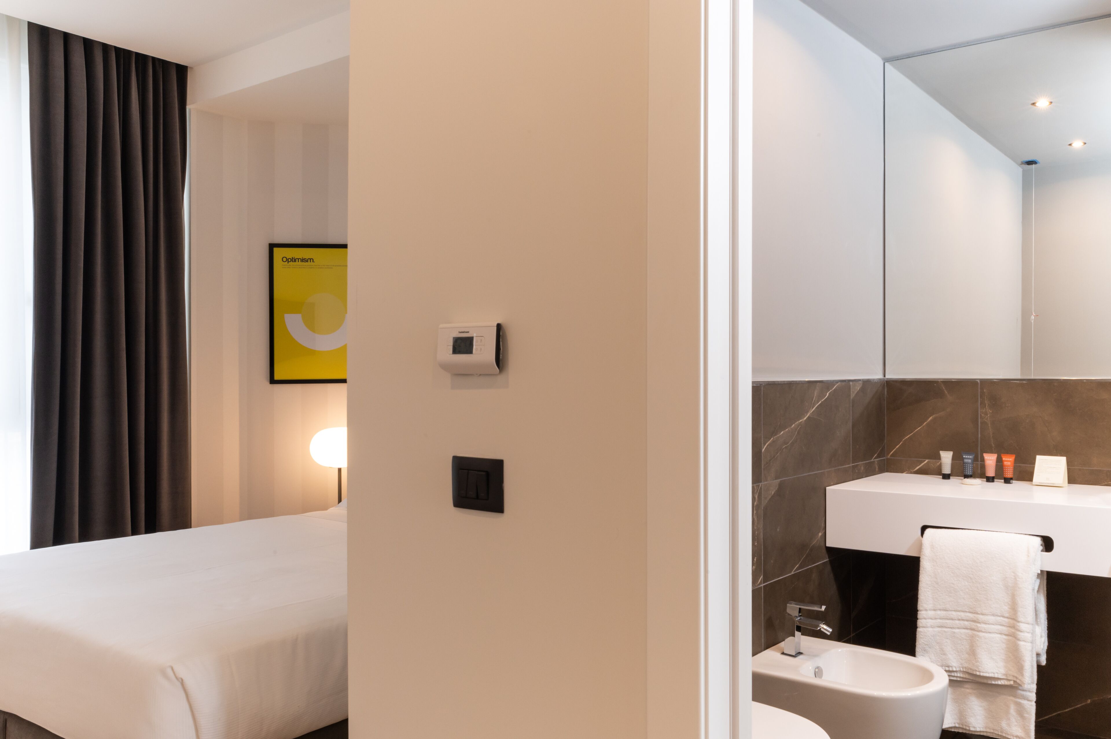 A modern hotel room with a bed, a bedside table, a wall-mounted thermostat, and a bathroom with a sink, mirror, and towel.