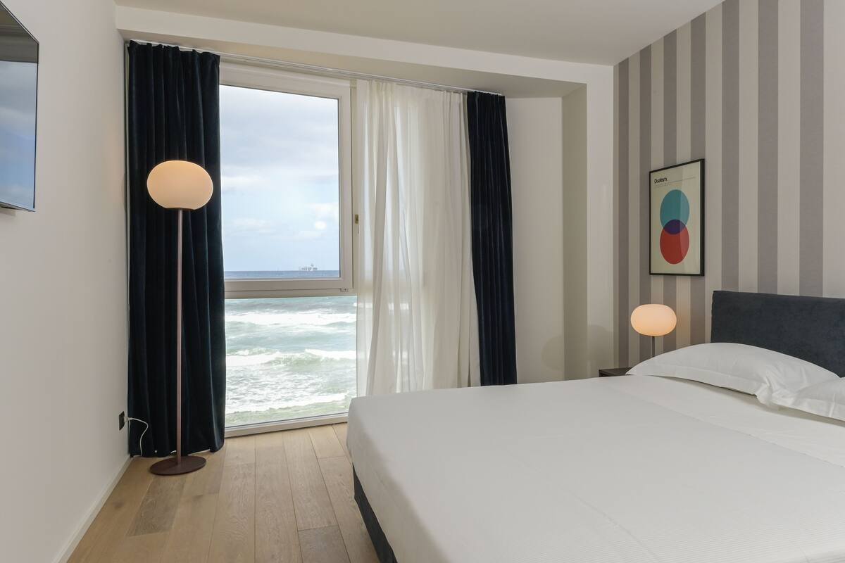 Une chambre d’hôtel avec un lit, une lampe sur pied, une grande fenêtre avec des rideaux et une œuvre d’art murale.