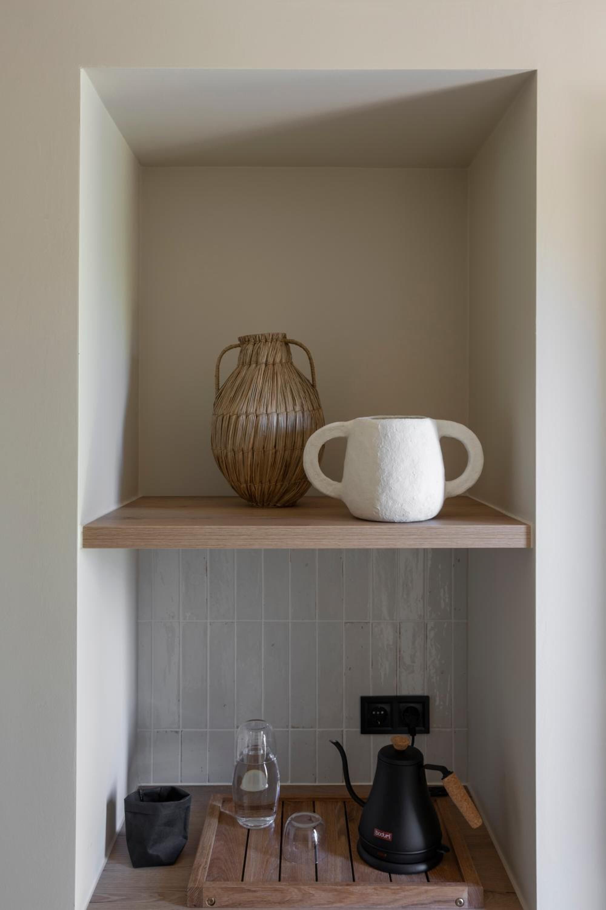 Een keuken met een houten plank waarop een geweven mand en een witte keramische mok staan. Daaronder een houten aanrecht met een waterkoker, een glas en een zwarte tas.