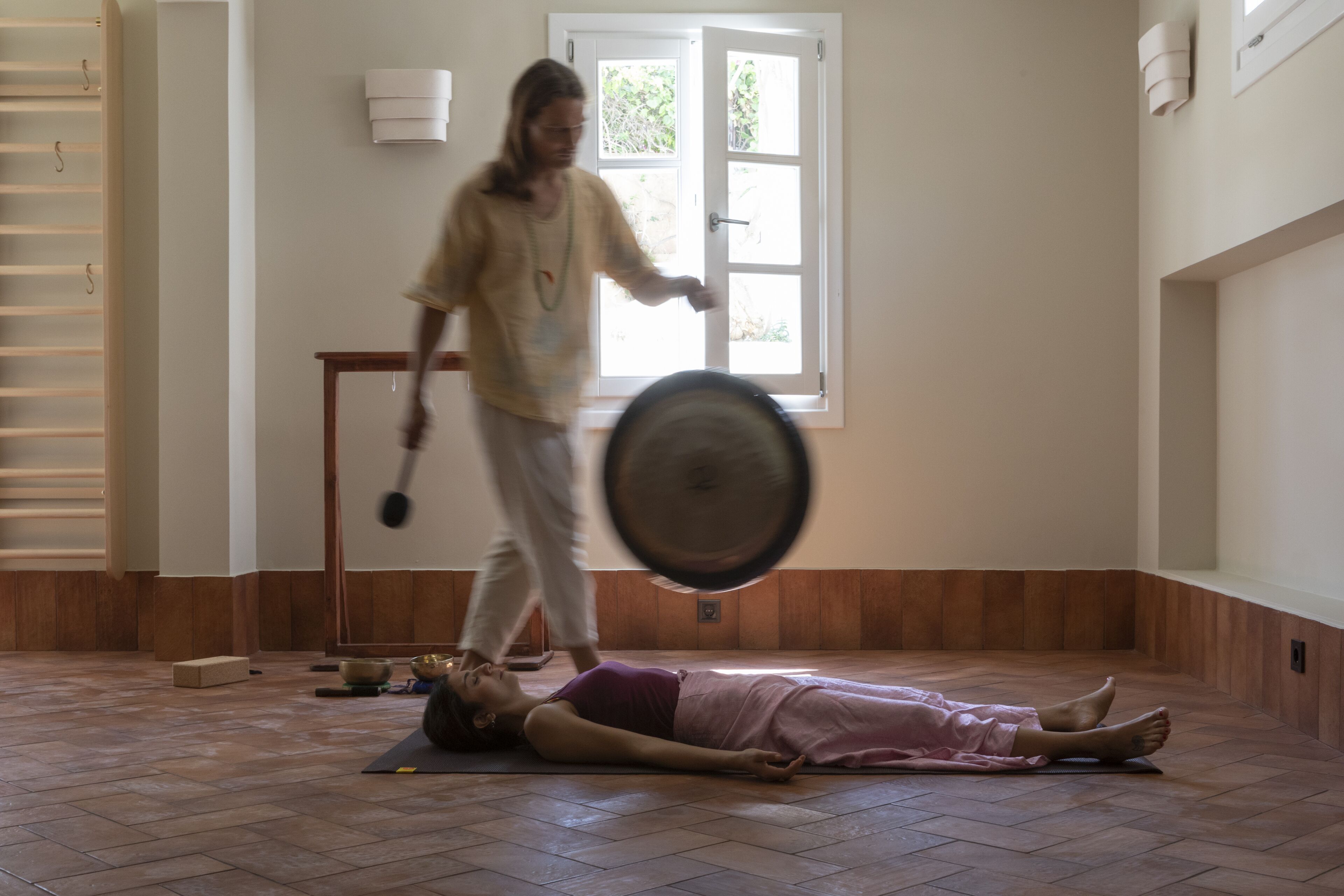 Una habitación con una persona acostada sobre una esterilla, otra persona sosteniendo un gong y una ventana que da a zonas verdes.