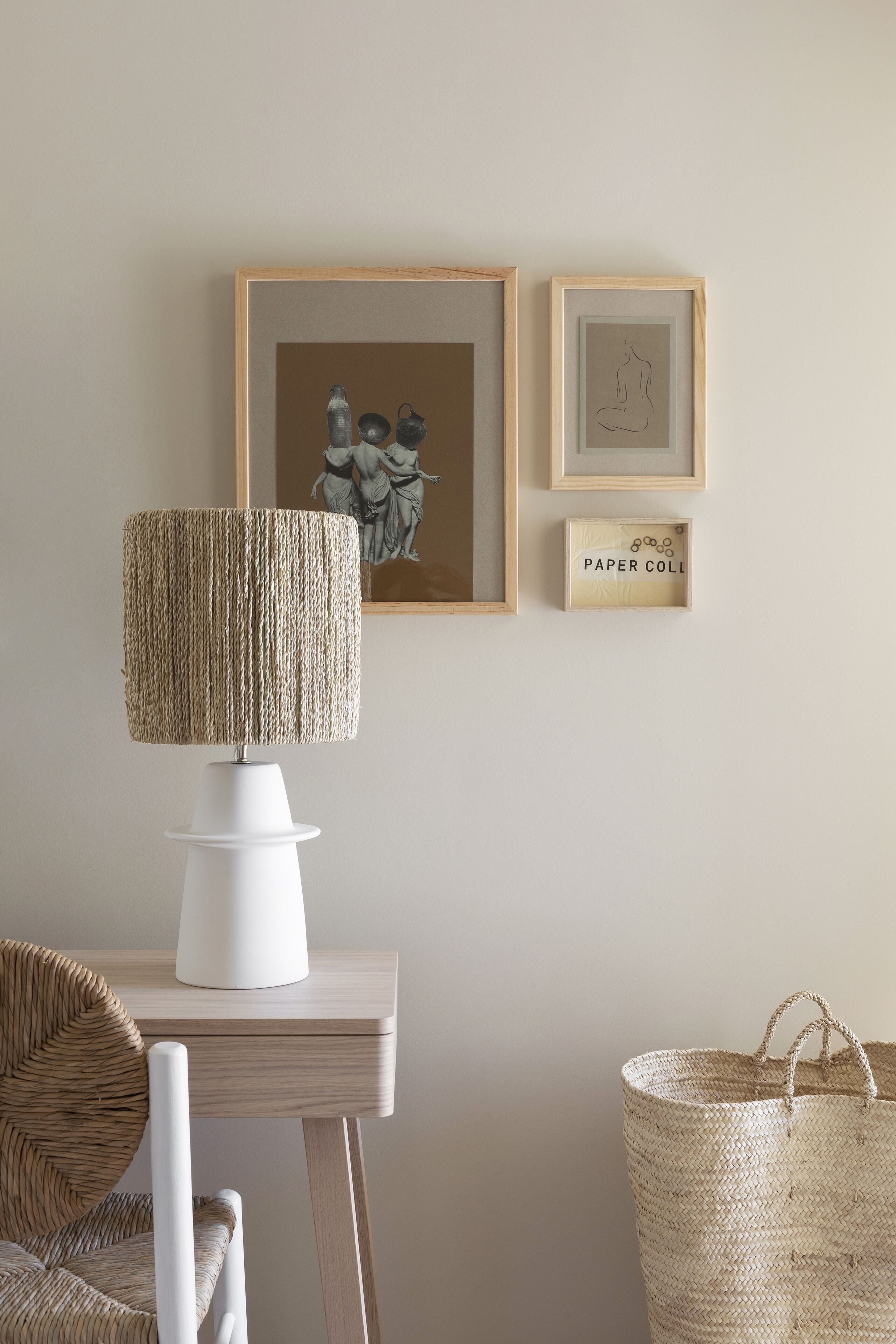 Una habitación con una lámpara de mesa blanca, una silla de mimbre, una cesta tejida y cuadros en la pared.