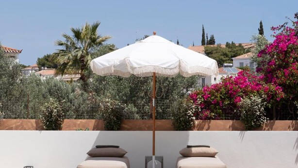 Un área junto a la piscina con una sombrilla blanca, dos sillones con cojines color beige y una terraza de madera.