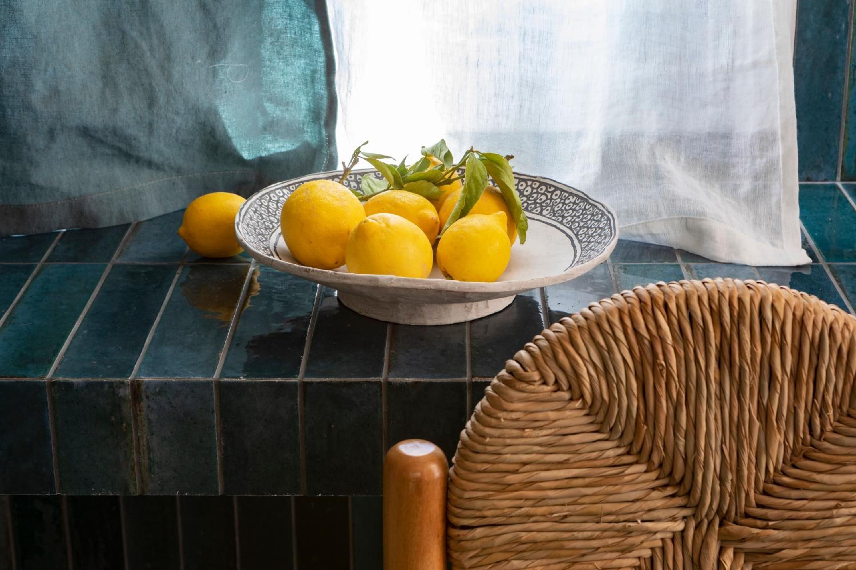Un cuenco de limones sobre una superficie de azulejos, con una cesta trenzada y una silla de madera.